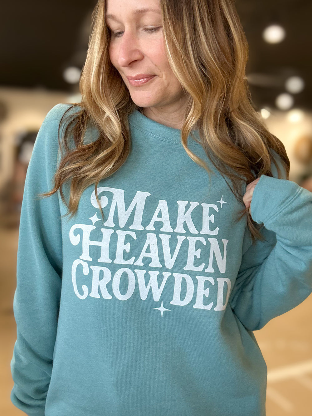 Woman wearing a teal sweatshirt with 'MAKE HEAVEN CROWDED' text.
